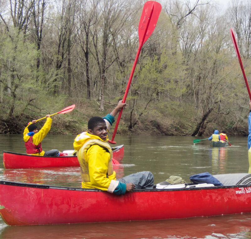 The image shows a group of people canoeing on a river. The person in the foreground is a young man in a yellow life vest, holding a red paddle. He is sitting in a red canoe. Other canoes with people are visible in the background, along with trees lining the riverbank. The water is calm, and the sky is overcast.